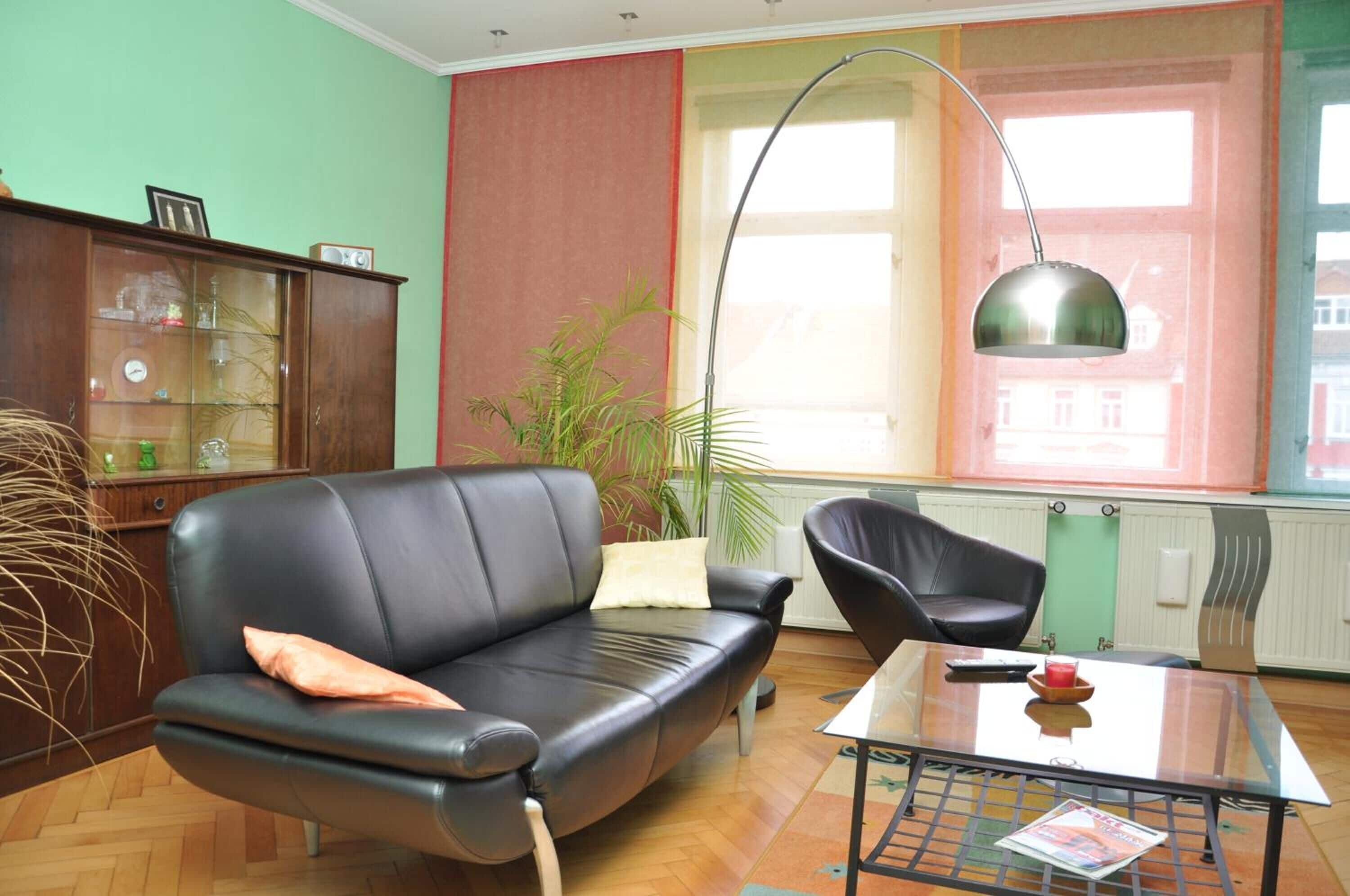 A living room with a black leather sofa, a glass coffee table, a floor lamp, a wooden cabinet, and a large window with curtains.