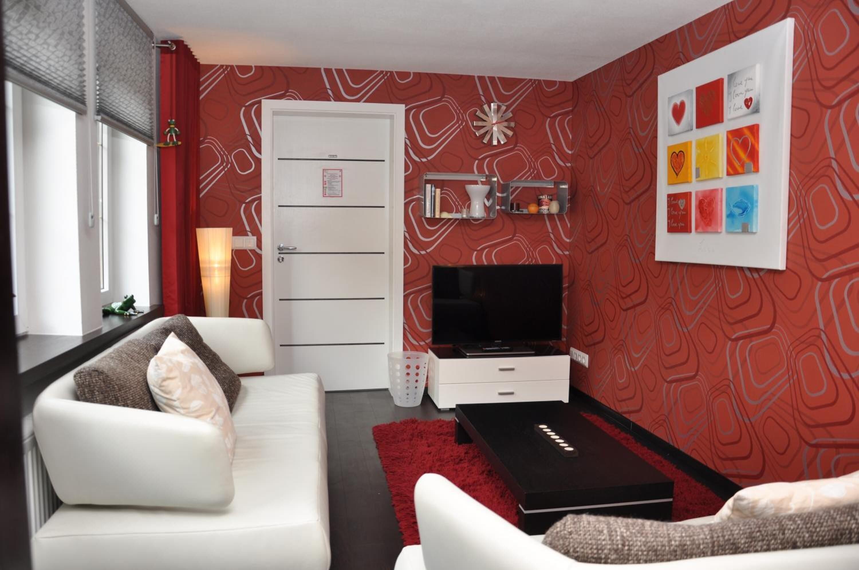 A modern living room with a white sofa, a black coffee table, and a red rug. There is a TV on a stand, a white cabinet, and a red patterned wall.