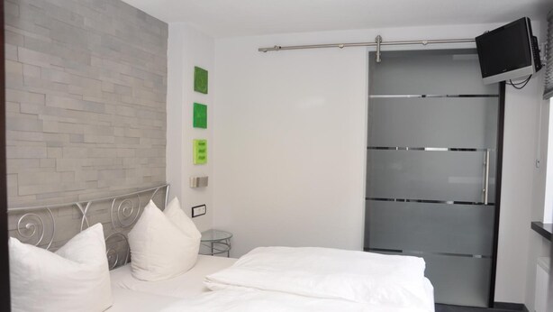 Une chambre d’hôtel avec un lit, du linge de lit blanc, une tête de lit ornée de motifs métalliques, une petite table de chevet, un téléviseur fixé au mur et une porte en verre dépoli.