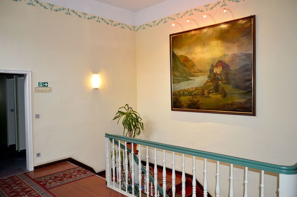 A hallway with a painting of a landscape, a staircase with a railing, and a potted plant.