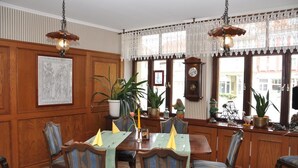 Un coin repas avec des murs lambrés, une grande fenêtre ornée de rideaux en dentelle et une table dressée pour quatre personnes, recouverte d’une nappe et de serviettes vertes.