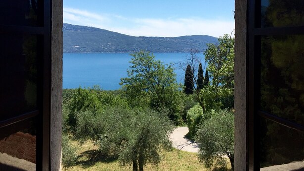 Vista desde una ventana a un lago rodeado de vegetación y colinas lejanas.