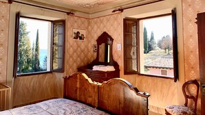 Un dormitorio con cama de madera, dos ventanas con vistas a árboles, un espejo y un aparador.