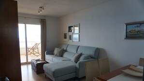 A modern living room with a light blue sofa, a wooden coffee table, and a dining table with chairs. There are framed pictures on the wall and a view of the outside through a sliding glass door.