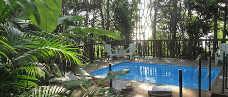 A swimming pool surrounded by a wooden deck, with white chairs and a covered seating area.
