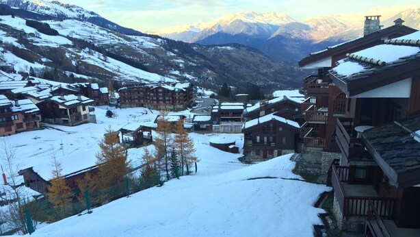 Un village enneigé, avec ses bâtiments traditionnels en bois, une piste de ski et des montagnes en arrière-plan.