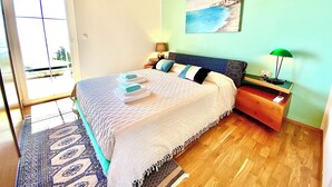 Un dormitorio con cama, mesita de noche, lámpara, un cuadro y vistas al exterior.
