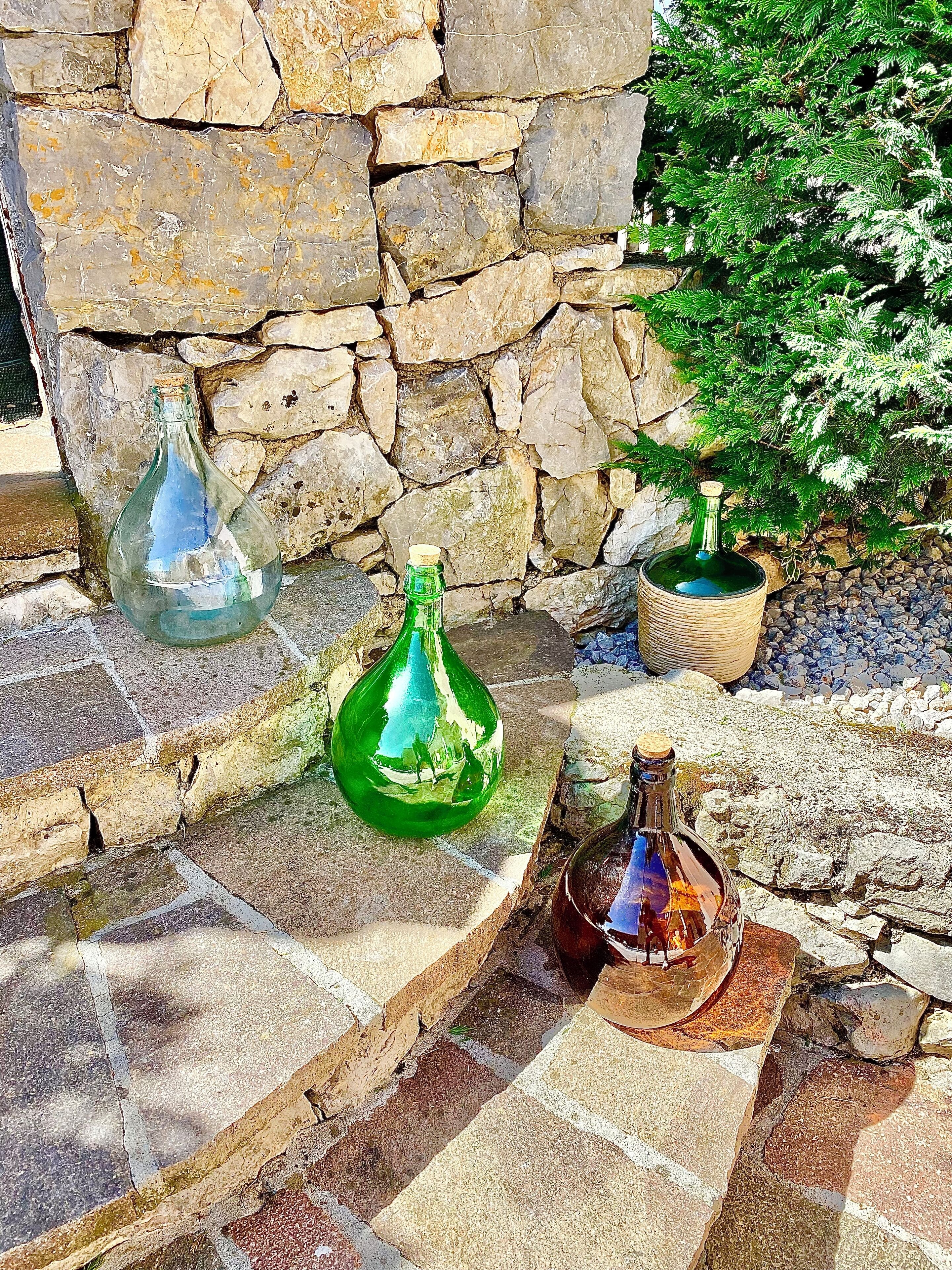 Trois bouteilles en verre sont posées sur une corniche en pierre, avec un mur de pierre et un petit arbre en arrière-plan.