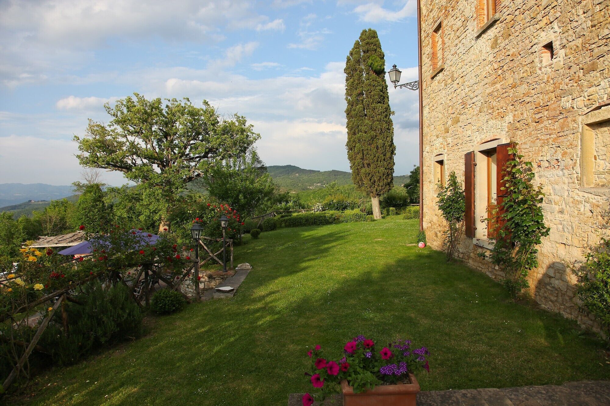 A stone building with a well-maintained garden, including a lawn, flowering plants, and a tall tree.