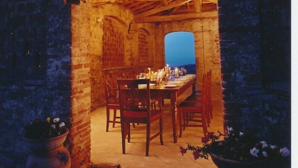 An arched stone room with a wooden table set for dining, surrounded by chairs, and a view of the night sky.