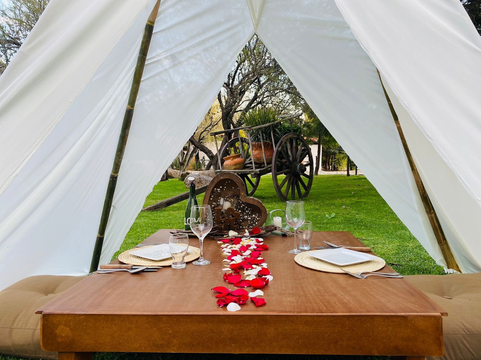 Une tente avec une table dressée pour deux personnes, comprenant une table en bois rustique, une nappe blanche et une brouette décorative en arrière-plan.