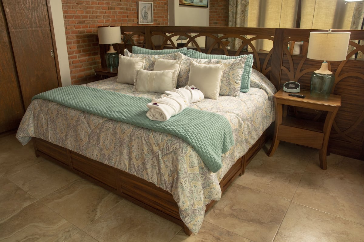 Una cama bien hecha con una colcha estampada, almohadas y una toalla doblada. Se aprecia un cabecero de madera, una mesita de noche con una lámpara y un cuadro colgado en la pared.