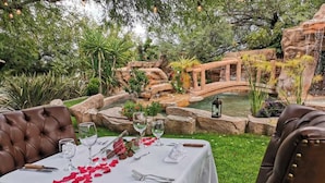 Uma área de refeições ao ar livre com uma mesa posta para dois, rodeada por vegetação exuberante e uma pequena cascata.