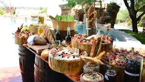 Uma mesa de buffet com diversos alimentos e bebidas, montada ao ar livre perto de uma piscina.