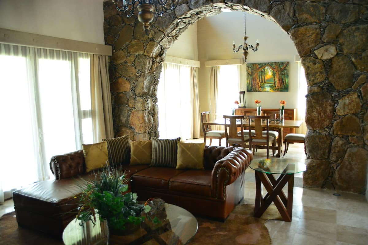 Una sala de estar con paredes de piedra, un sofá de cuero, una mesa de centro de vidrio y una zona de comedor con una mesa y sillas de madera.