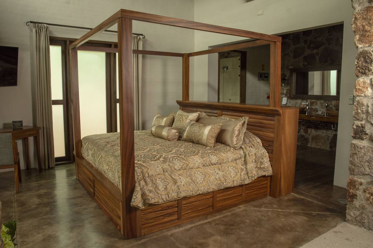 Una cama con dosel, colcha estampada y varias almohadas, una mesita de noche de madera y una pared de piedra al fondo.