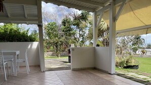 A covered patio area with tiled flooring, a white table and chairs, and a view of a garden with various plants and a lawn. - Apartment/ flat - Petit-bourg (PETIT BOURG)