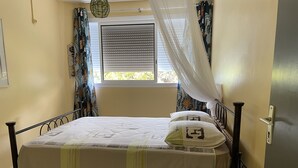 A bed with a white bedspread and pillows, a small bedside table with a remote control, a window with blinds, and a curtain with a leaf pattern. - Apartment/ flat - Petit-bourg (PETIT BOURG)