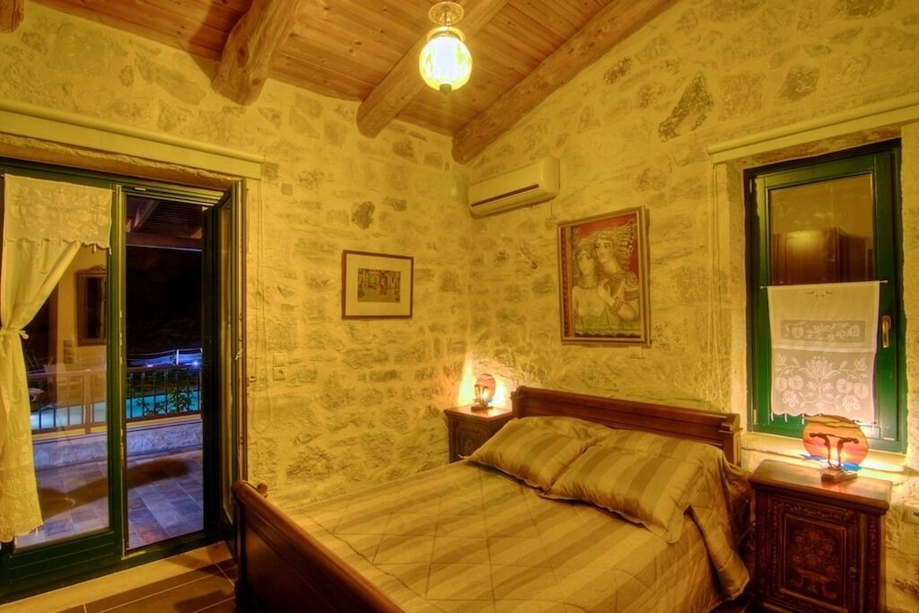 Une chambre avec un plafond en bois, des murs en pierre, un lit recouvert d’une courtepointe, une table de chevet avec une lampe et une fenêtre dotée d’un cadre vert.