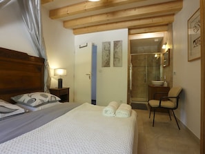Un dormitori amb llit, cadira, tauleta de nit, bany amb dutxa i porta que dona a una altra habitació.