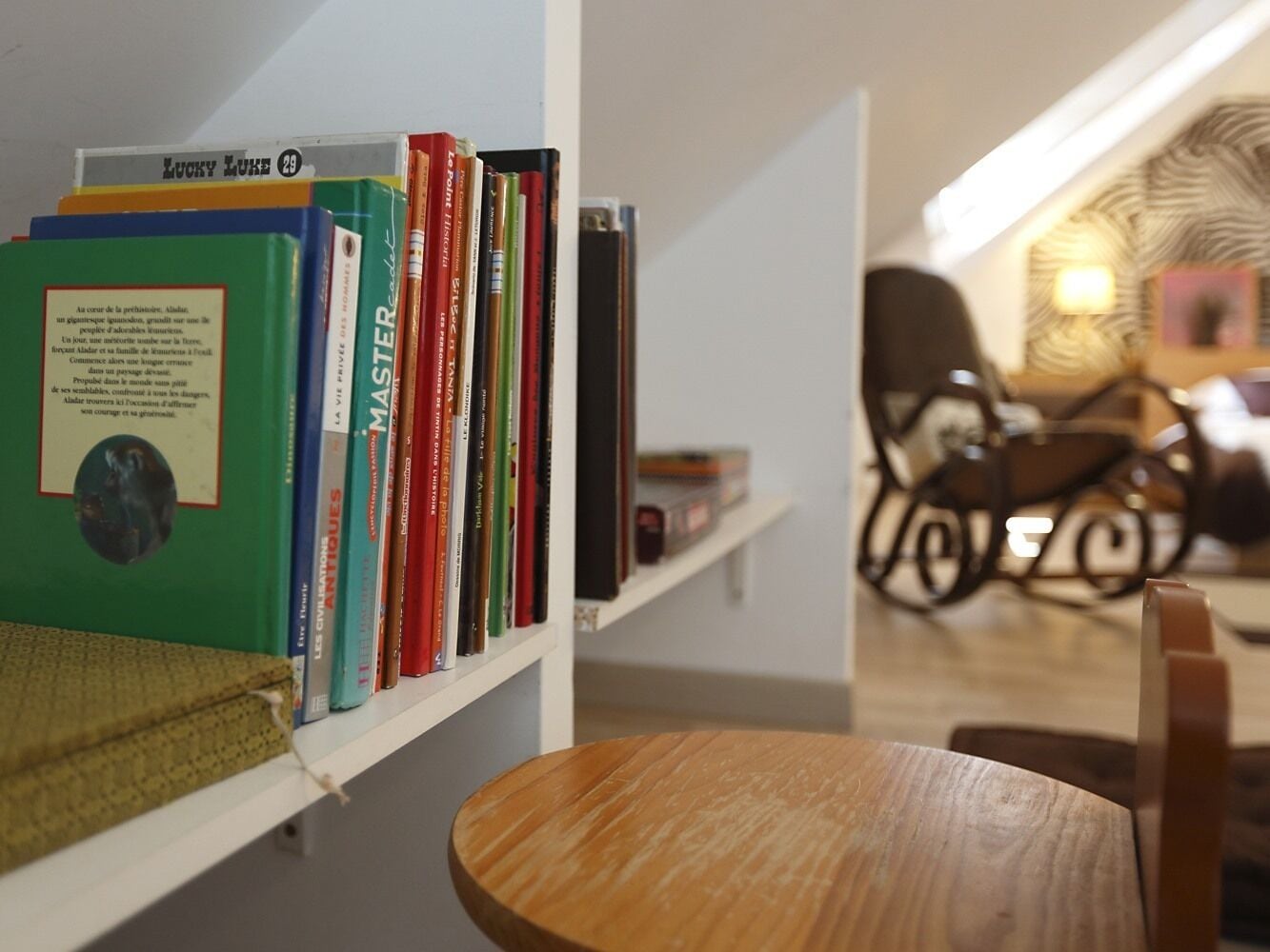 A bookshelf with various books and a small round table in a room with a chair and a lamp in the background.