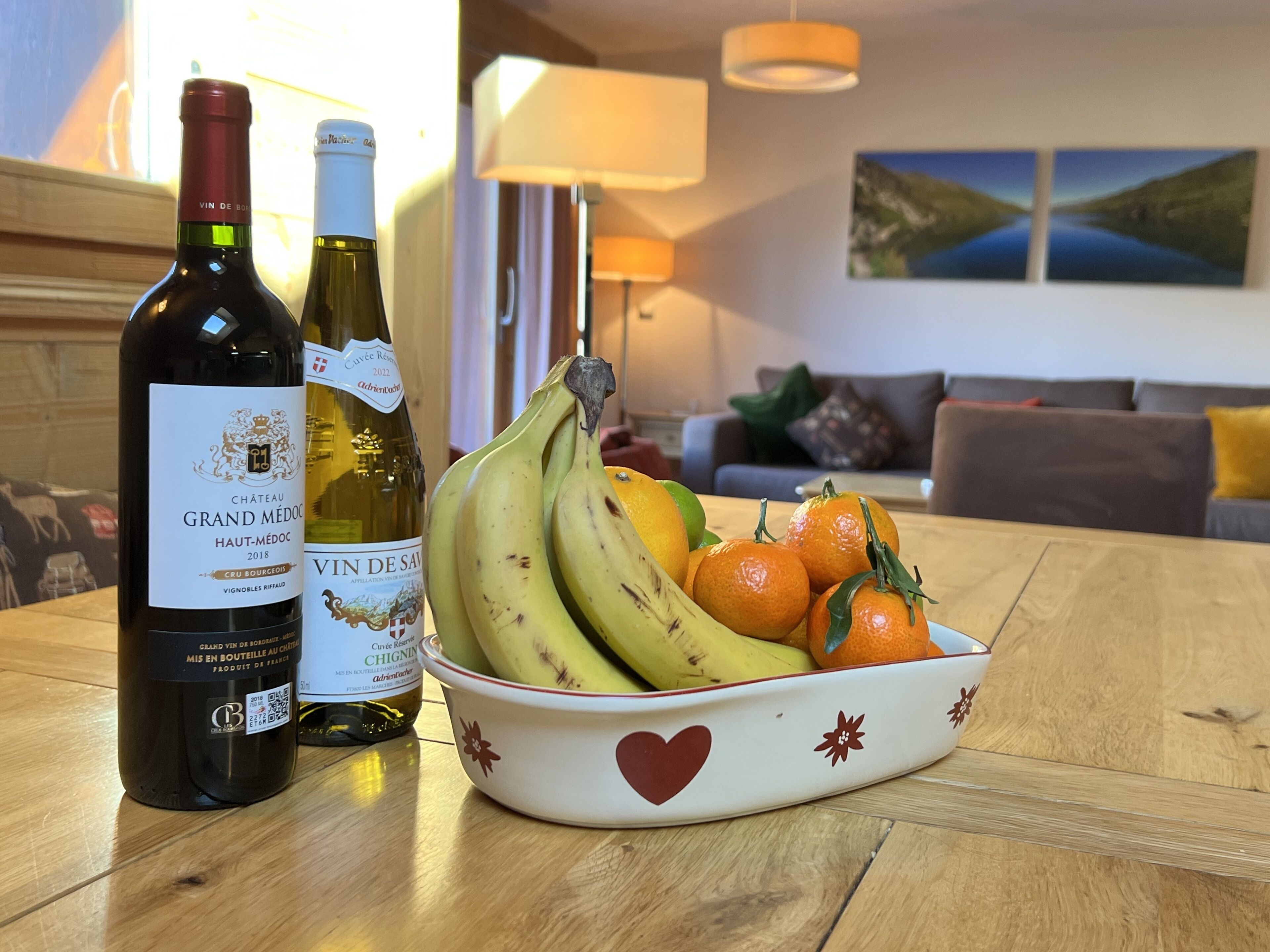 Twee flessen wijn op een houten tafel, een fruitschaal en een woonkamer op de achtergrond.