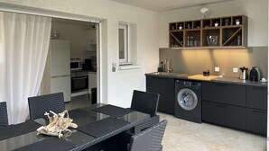 A compact kitchen with a washing machine, a dining area with a table and chairs, and a small shelf with kitchen items.