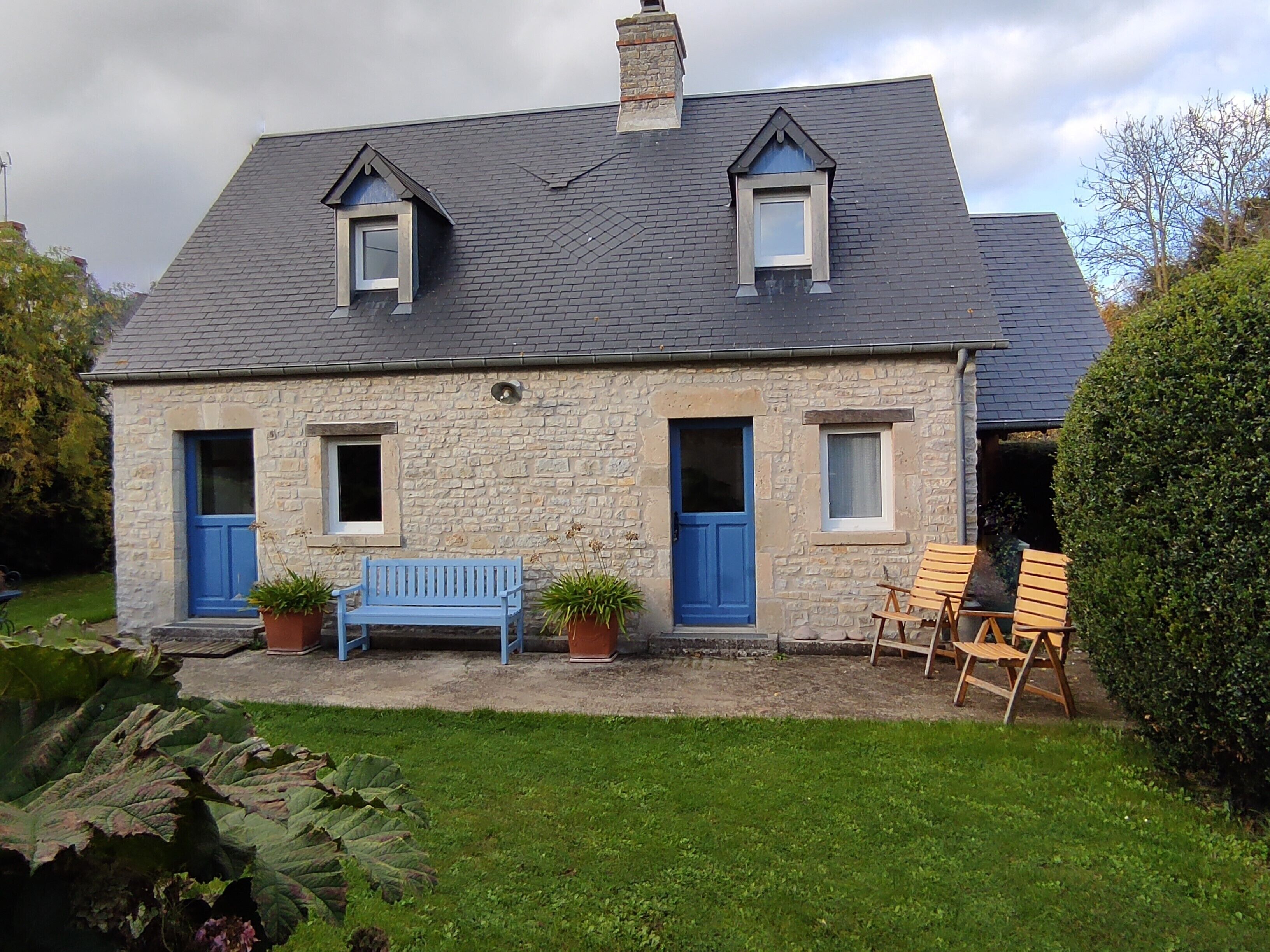 A stone house with a sloped roof, two dormer windows, and a front porch with a blue bench and wooden chairs.
