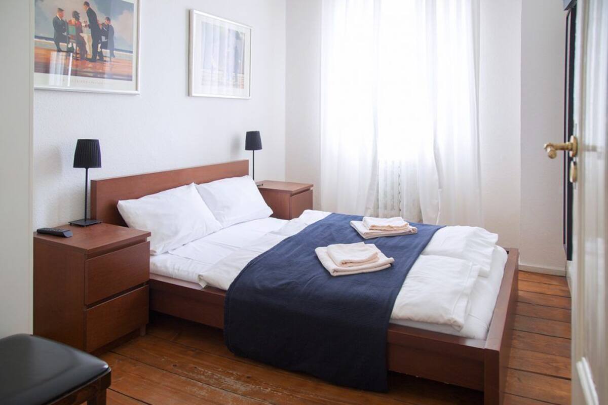 A neatly made bed with white linens and a blue blanket, two bedside tables with lamps, a wooden floor, and a door to the right.