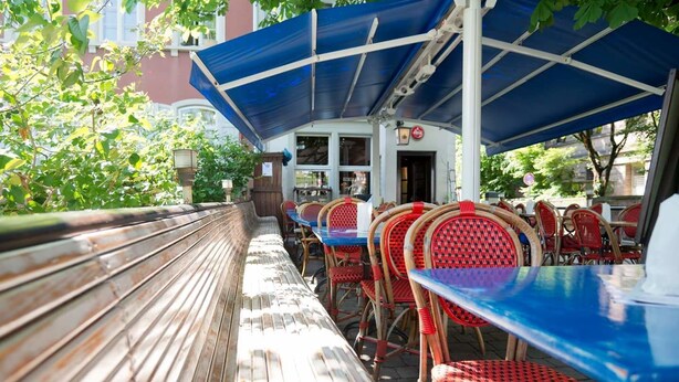 An outdoor seating area with wooden benches and red wicker chairs under a blue canopy. - Hemingway's Hostel Heidelberg (Heidelberg)
