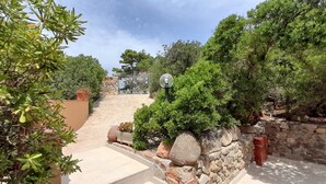 Un chemin pavé mène à une entrée fermée par un portail, qui est elle-même entourée d’une végétation luxuriante et de murs en pierre.
