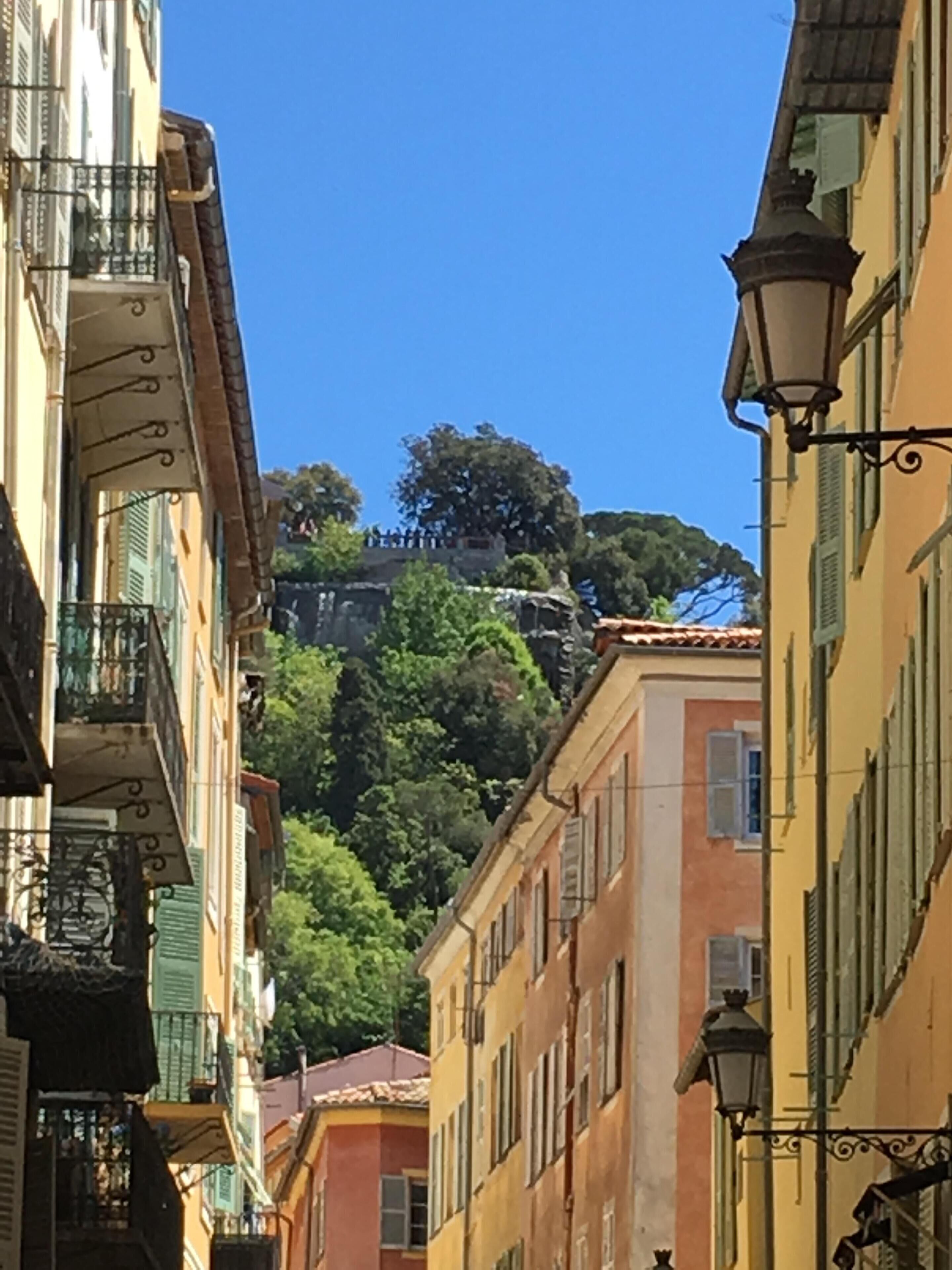A narrow street with buildings on either side, featuring balconies and a street lamp.