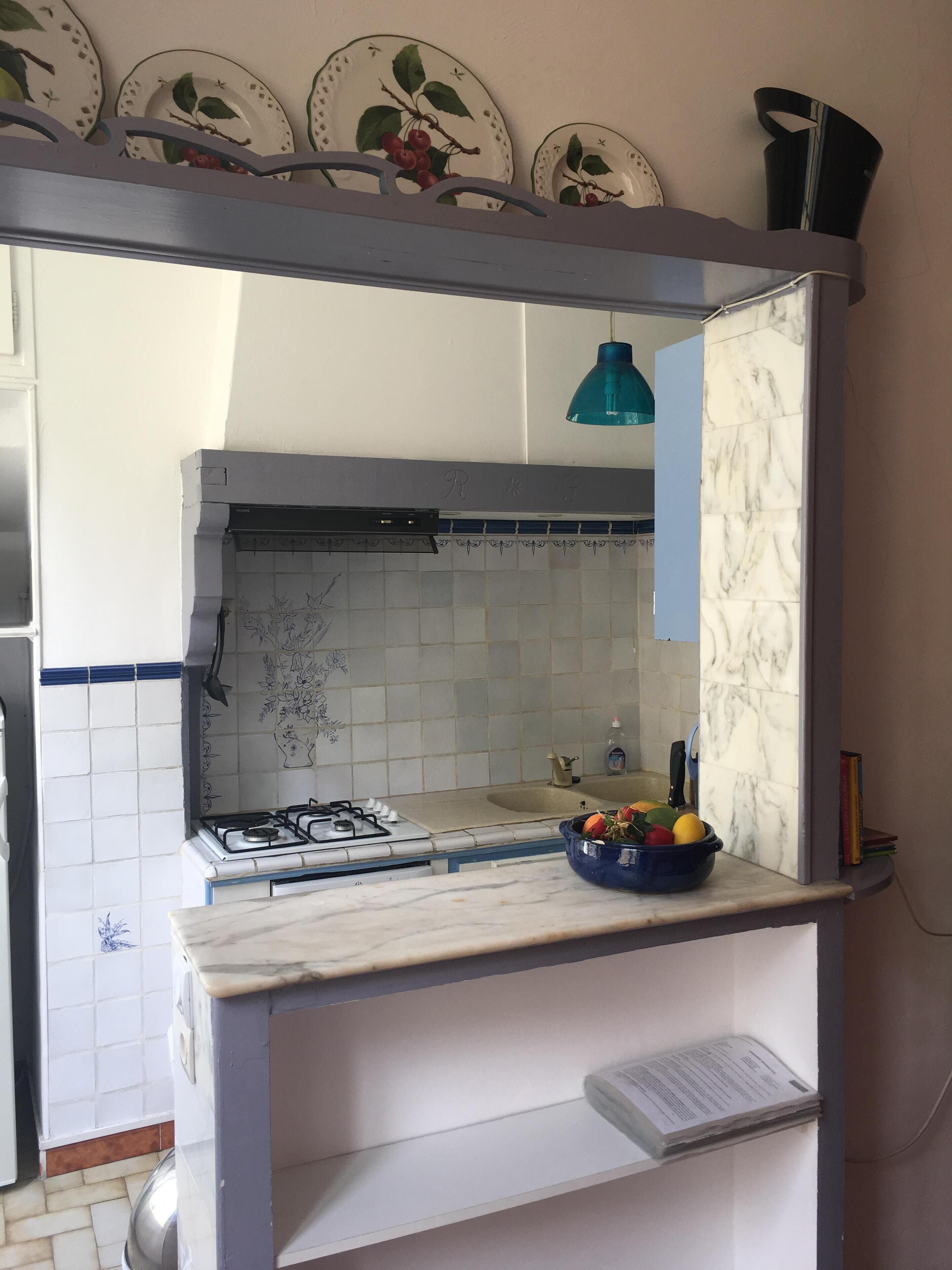 Una cocina compacta con estufa a gas, una lámpara colgante azul y un cuenco de fruta en la encimera.