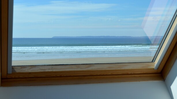 A view through a window of a beach with waves, a clear sky, and a distant landmass. - Duplex apartment with large terrace facing the sea (plomodiern)