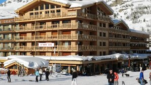Un bâtiment en bois à plusieurs étages, doté de balcons, d’un local pour le matériel de ski et d’une enseigne sur laquelle on peut lire : « C’est ici ».