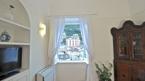 Ein Zimmer mit Fernseher auf einer weißen Kommode, einem Fenster mit weißen Vorhängen, einer Topfpflanze und einer Holzkabine mit Glastüren.