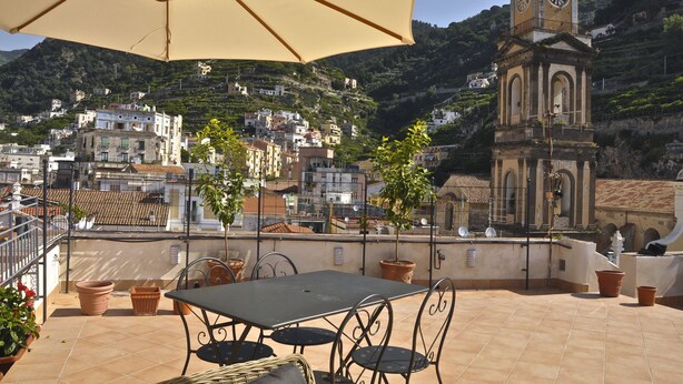 Eine Dachterrasse mit Tisch und Stühlen, einem großen Sonnenschirm und einem Kirchturm im Hintergrund.
