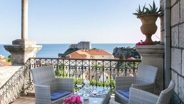Un balcón con sillas de mimbre, una mesa de vidrio y vistas al mar y edificios.
