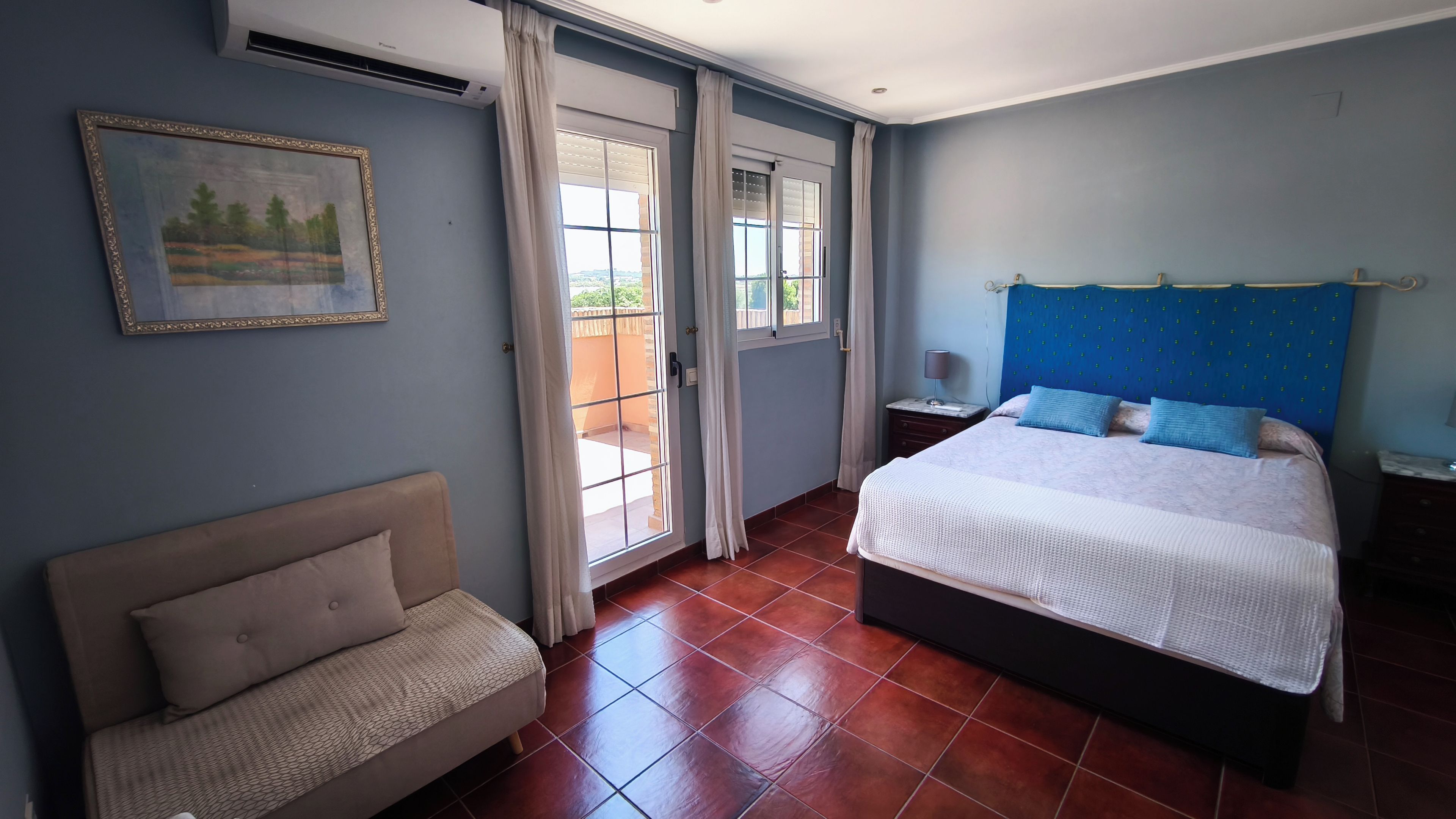 Un dormitorio con cama, sofá, mesita de noche, lámpara y ventana con cortinas.
