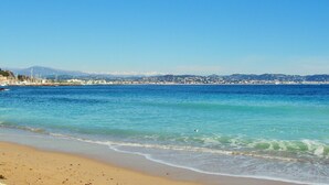 A beach with clear blue water, a sandy shore, and a distant cityscape with buildings and hills. - The calm of the forest, just minutes from the sea (Fréjus)