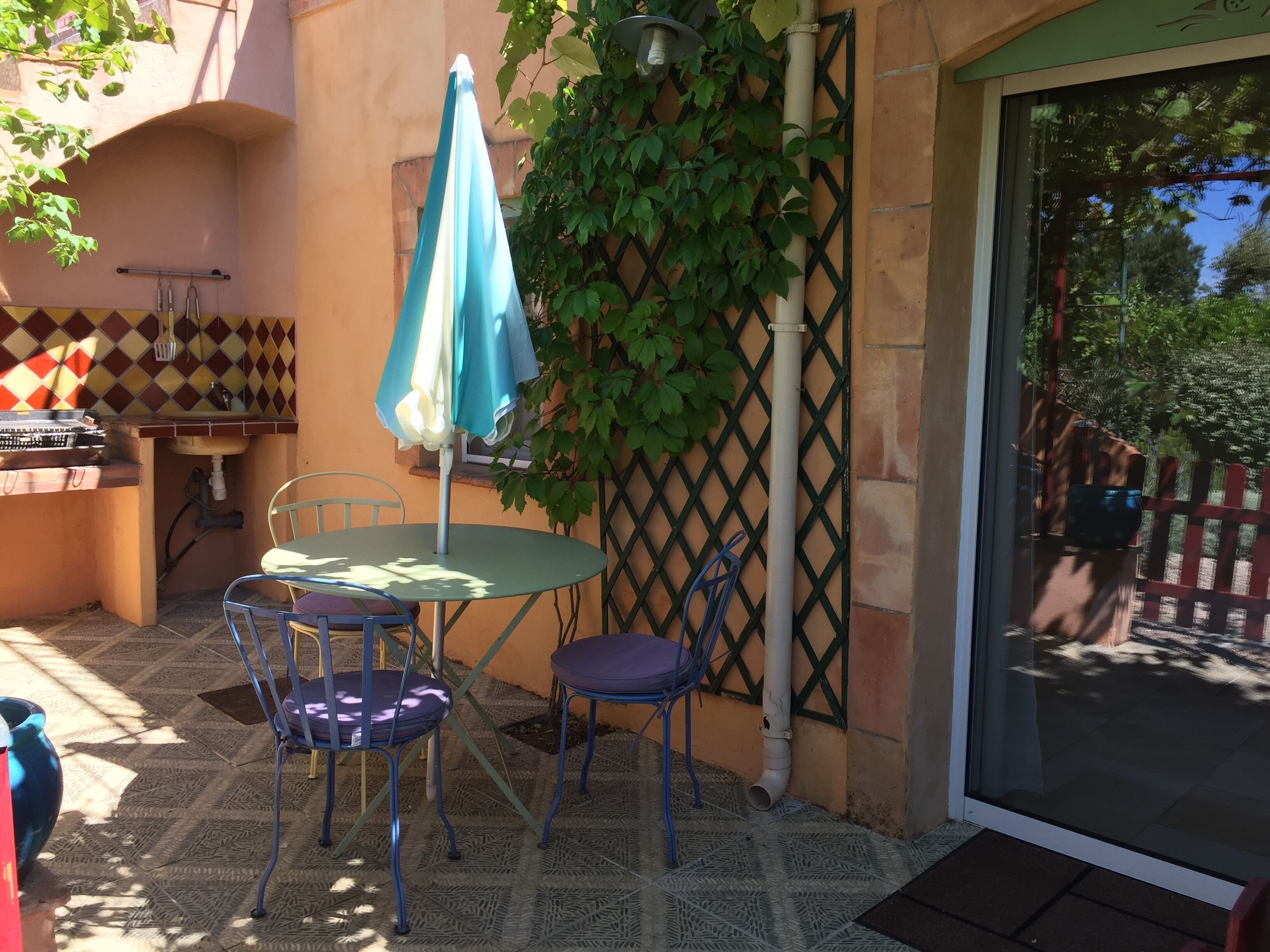 Un espace patio aménagé avec une table et des chaises, un parasol bleu, un évier et un mur recouvert de plantes grimpantes.