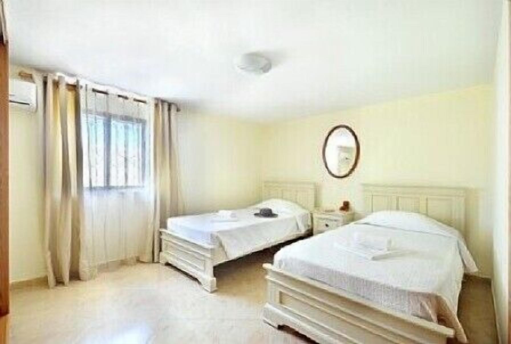 A hotel room with two single beds, a small bedside table with a lamp, a window with curtains, and a round mirror on the wall.