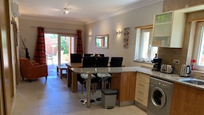 Een moderne keuken met een ingebouwde wasmachine, een eethoek met tafel en stoelen, en een woonkamer met een bank en gordijnen.