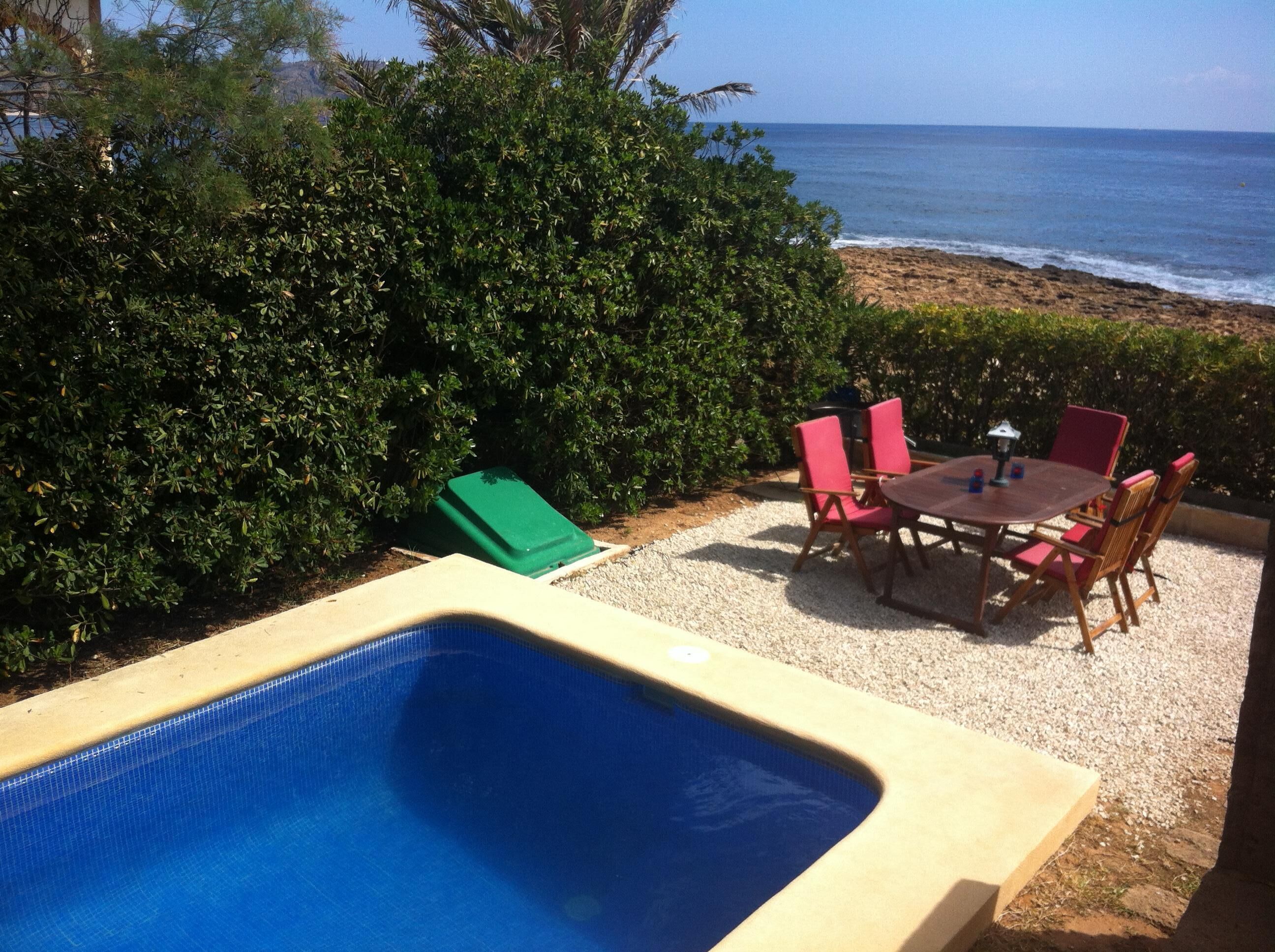A pool with a table and chairs, surrounded by a hedge and a view of the ocean.