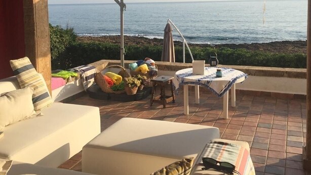 A cozy outdoor seating area with a view of the sea, a table with a tablecloth, and a white umbrella providing shade.