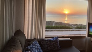 A room with a view of the sunset over the sea, a sofa with patterned cushions, and a television on a stand.