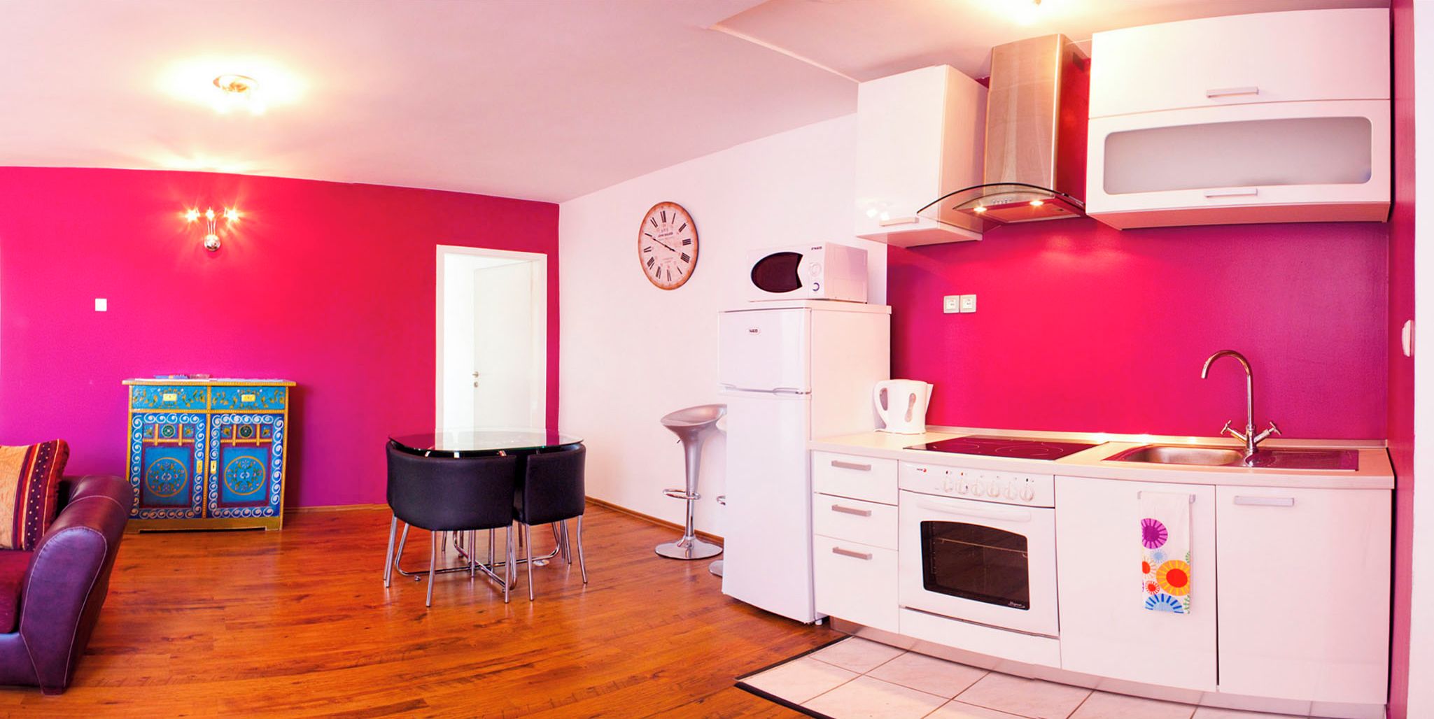 Une cuisine compacte avec des appareils électroménagers blancs, un coin repas doté d’une table en verre et de chaises noires, et un mur d’un rose vif.