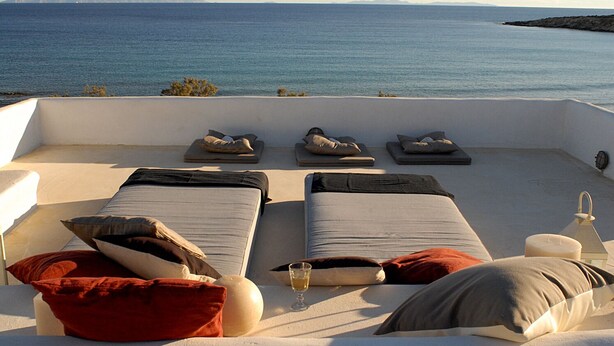 Een zonnig terras met twee bedden, kussens, een glas wijn en uitzicht op zee.