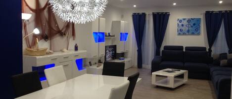 Uma sala de estar moderna com uma mesa de jantar branca, cadeiras pretas, um sofá azul e um grande lustre decorativo.