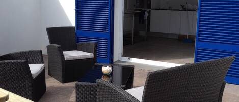 Um pátio externo com móveis de vime, uma mesa de madeira e vista do interior através de persianas azuis.